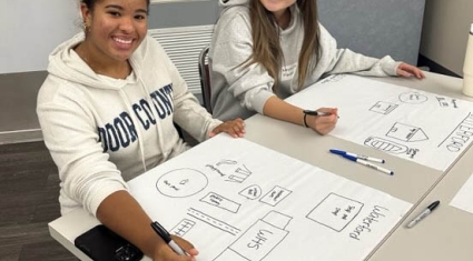 Two Student United Way participants seated at a table smiling at the camera as they work on community maps